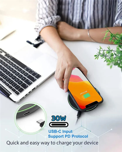 Person using a wireless charger with a smartphone, laptop, and plant on a desk.